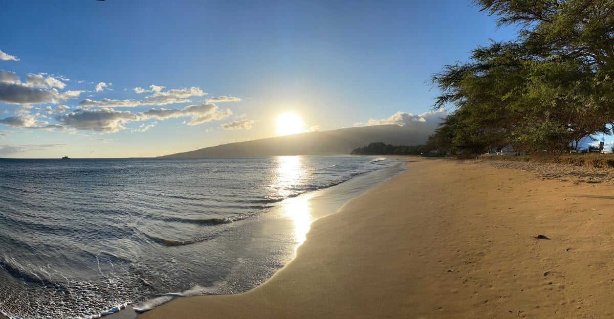 Scenic beach at sunset with golden sand, gentle waves, and a large mountain silhouetted against the bright sun on the horizon